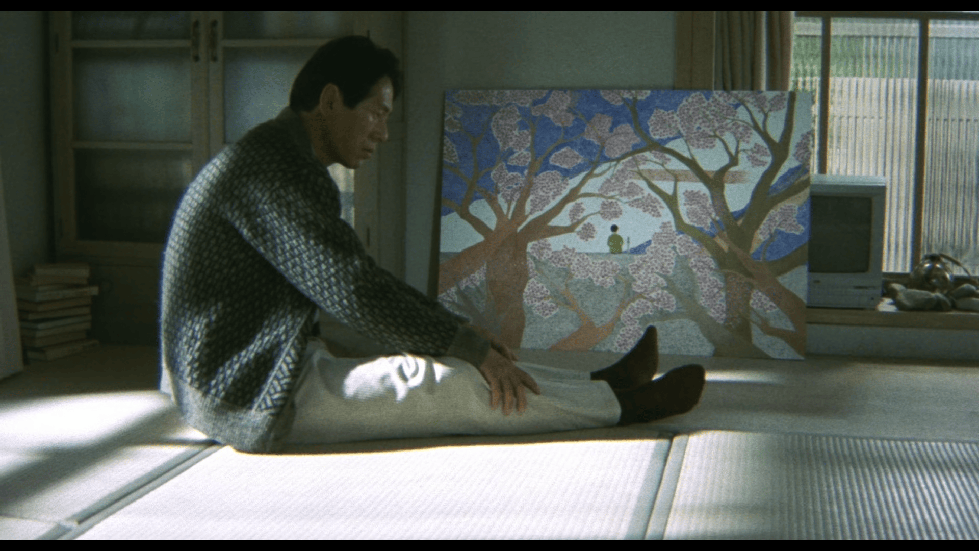 Shot from 1997's Hana-bi. A man sits slouched in  a tatami floor, holding his extended legs. Behind him is one of his paintings in the floor against the wall. The painting shows a myriad cherry blosssom trees framing a man in a kimono, sitting on his legs beside a blade that is stuck on the floor